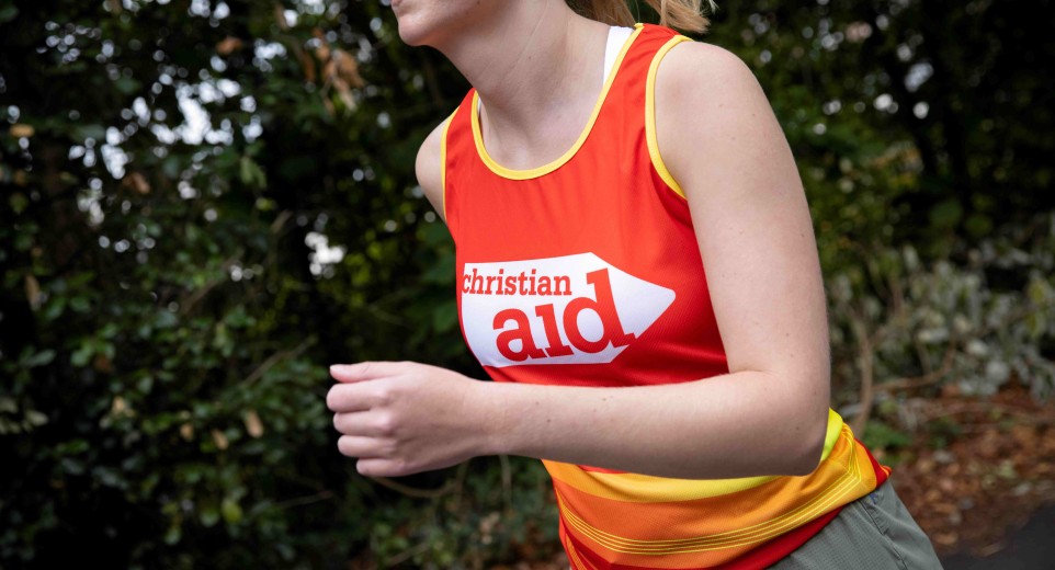 A woman running. There are trees behind her, and she's wearing and red and yellow Christian Aid running vest.