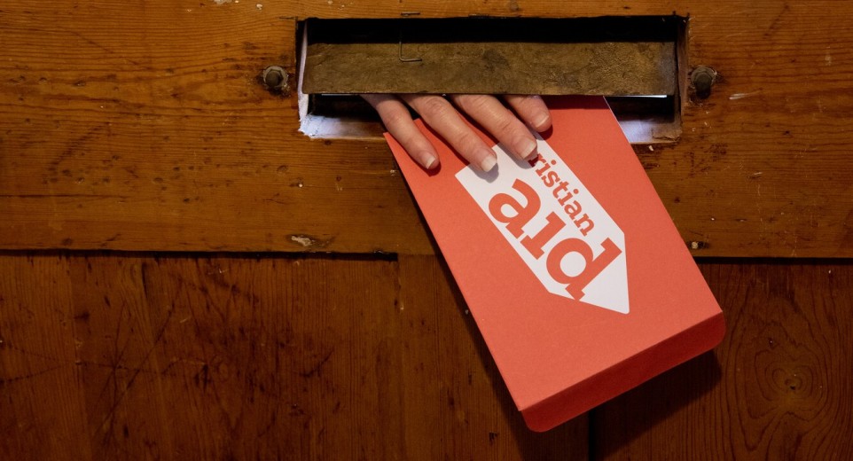 A hand reaching through the letterbox, holding a red Christian Aid envelope.
