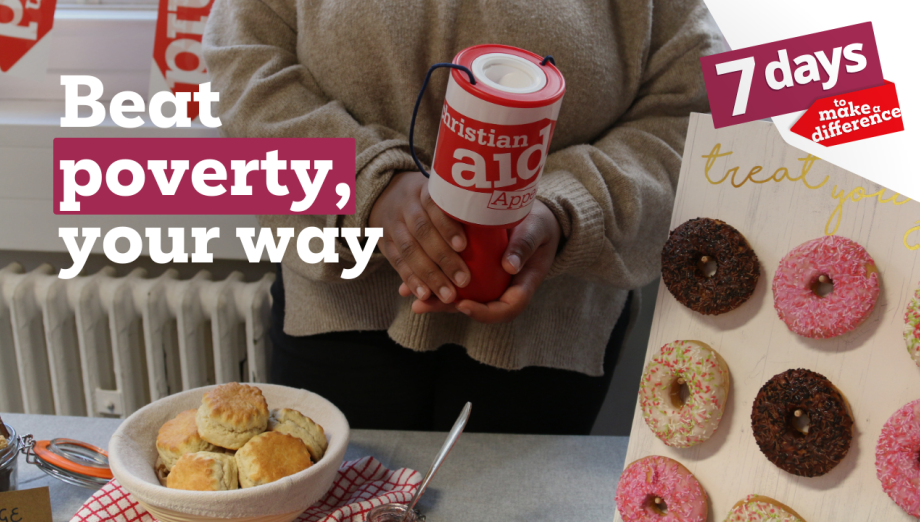 A person in a beige jumper holds a red Christian Aid donation tin at a fundraising event. In the foreground, a table displays a bowl of scones with a jar of jam and a spoon, alongside a stand with decorated doughnuts. Red Christian Aid bunting hangs in the background.