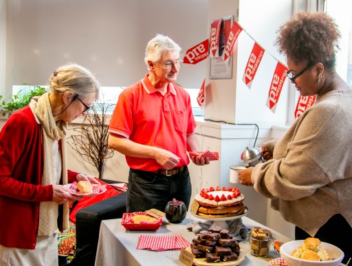 Two women and a man stood together. One woman is pouring tea for the man, whilst the other is putting cake on her plate. They are stood around a table full of baked goods.