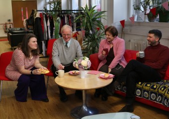 Four people sit on red chairs and sofas around a coffee table, smiling and chatting over tea and cakes in a cosy indoor setting decorated with bunting and clothing racks in the background.